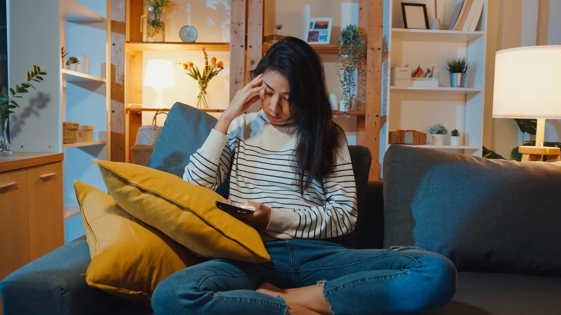 Thoughtful woman holding phone feeling sad waiting for call sit at sofa in living room at house night feel lonely.